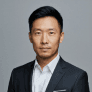 An Asian man wearing a formal suit and tie, standing confidently with a neutral expression.